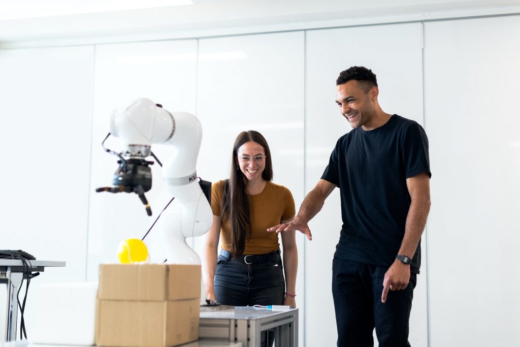 Zwei Menschen stehen lachend und miteinander interagierend vor einer Robotik Einheit.