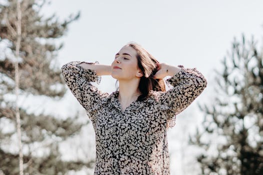 A woman stands outdoors, smiling with closed eyes, in a floral blouse, enjoying the serene environment.
